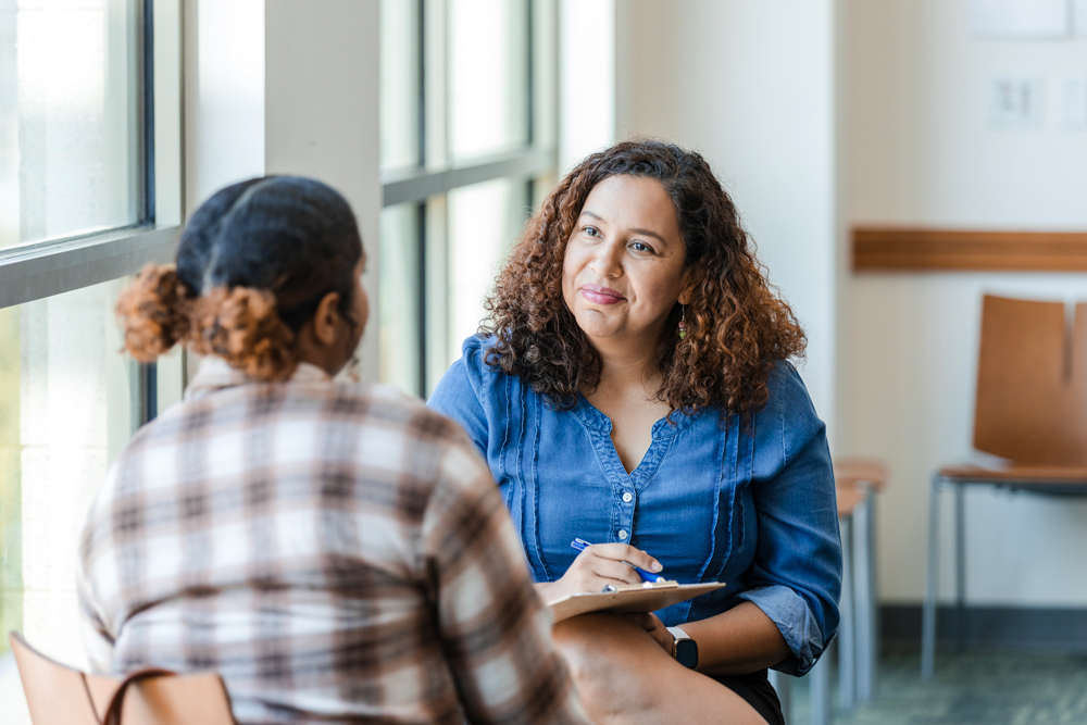 A woman converses with another woman in a waiting room, both appearing engaged and attentive to each other.