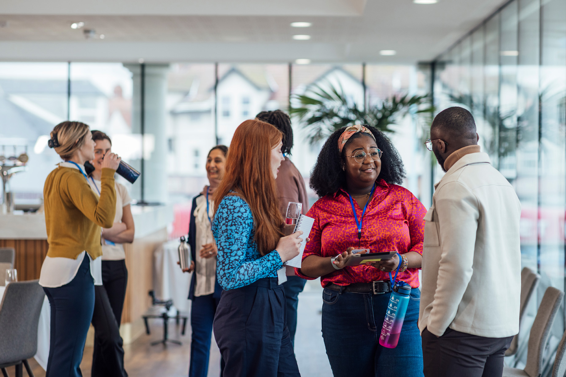 A diverse group of professionals wearing name badges talk and network in a bright, modern indoor space, holding notebooks and water bottles during a conference or workshop break.