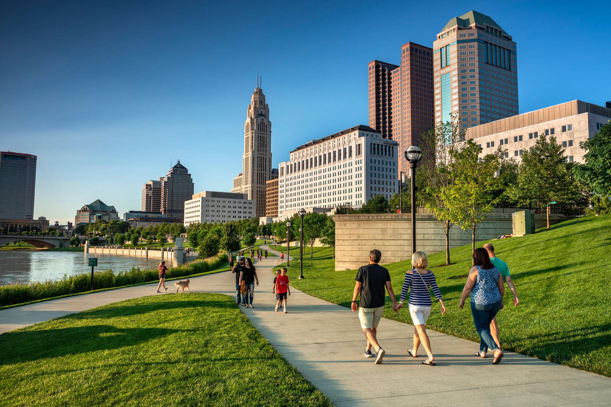 People stroll along a riverside path with city buildings visible in the background.