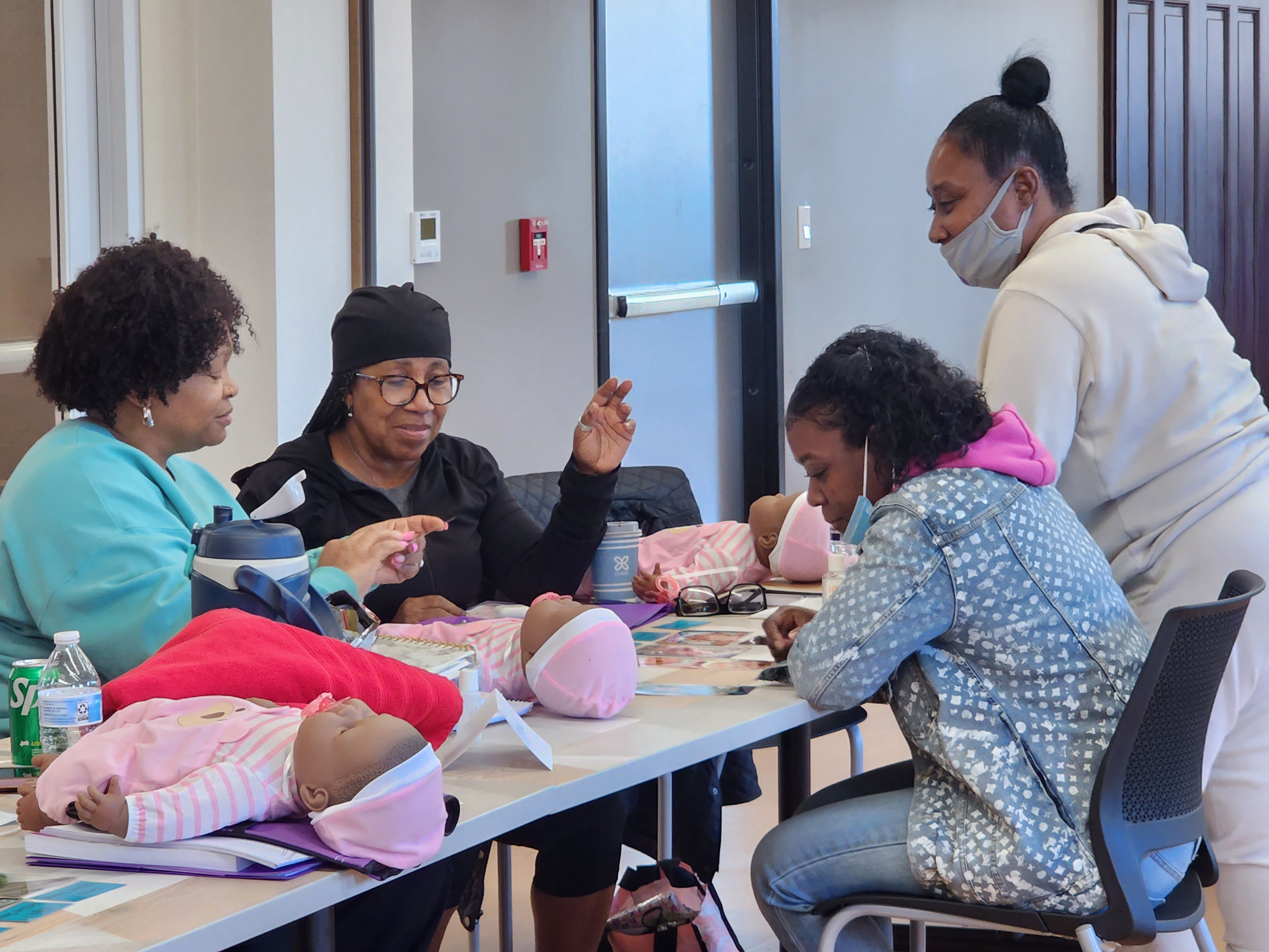Group of women seated around a table in a classroom, practicing infant care using baby dolls, with one woman demonstrating while others observe and participate.