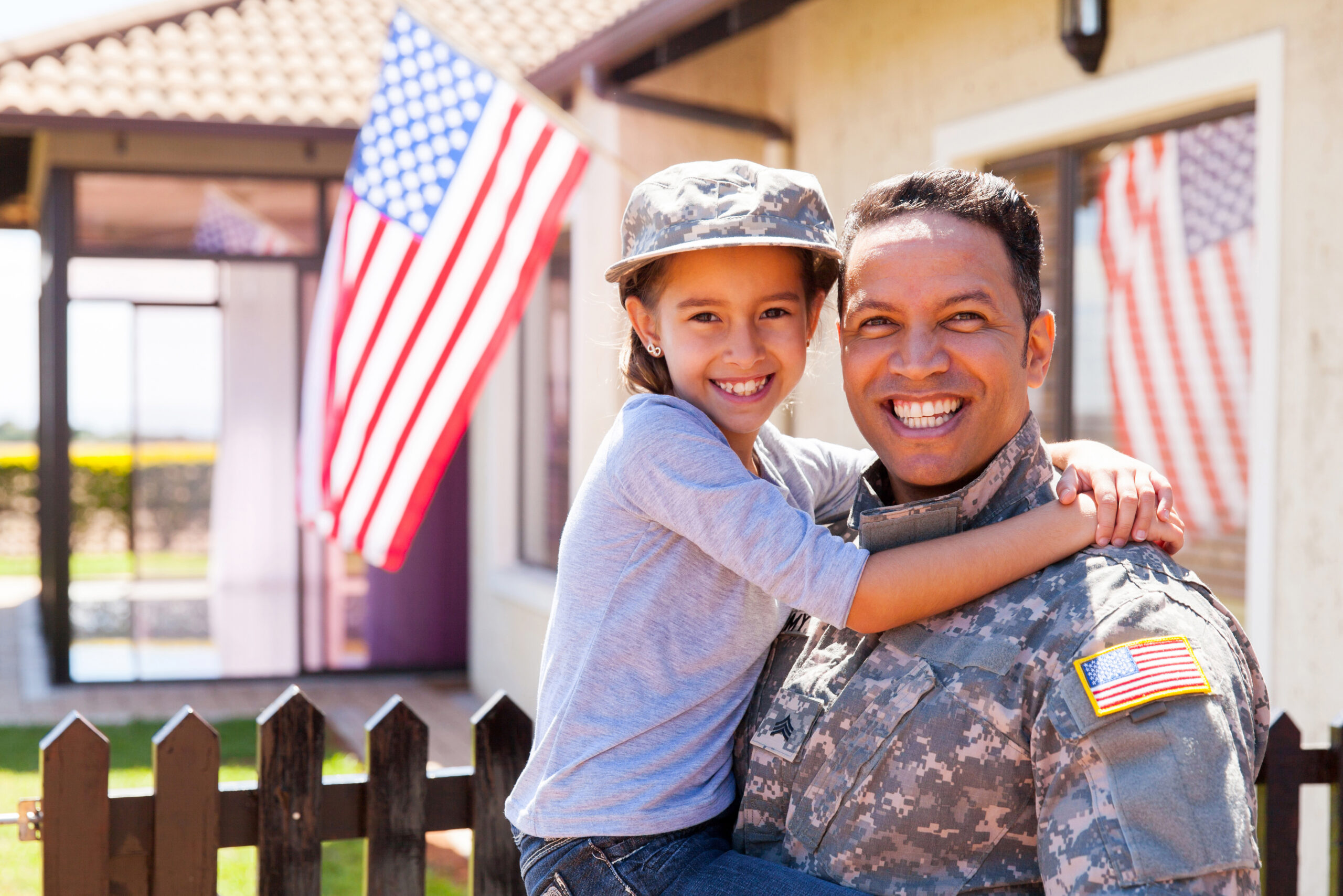 A smiling military service member in uniform holds a young child in front of a home with American flags, representing family support, veterans, service, and patriotism.