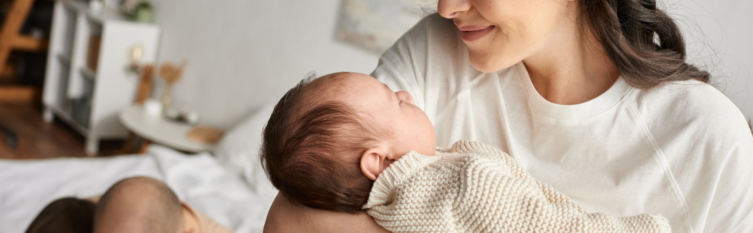 Mother holding and gazing at a newborn baby wrapped in a knit blanket in a softly lit bedroom.